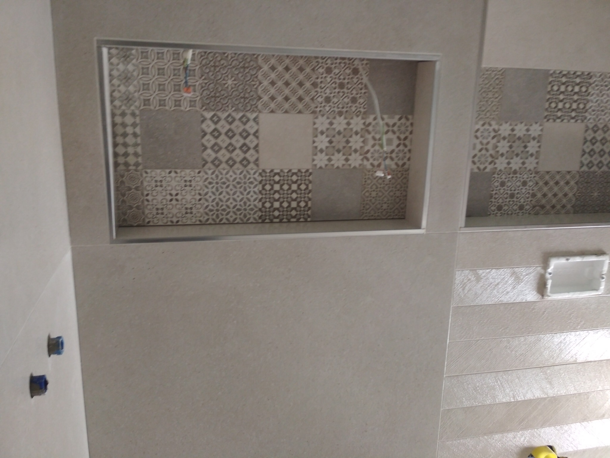 Beżowe płytki ścienne z dekoracyjnymi wnękami wyłożonymi mozaiką w łazience, widoczne przyłącza wodne i otwór na oświetlenie.