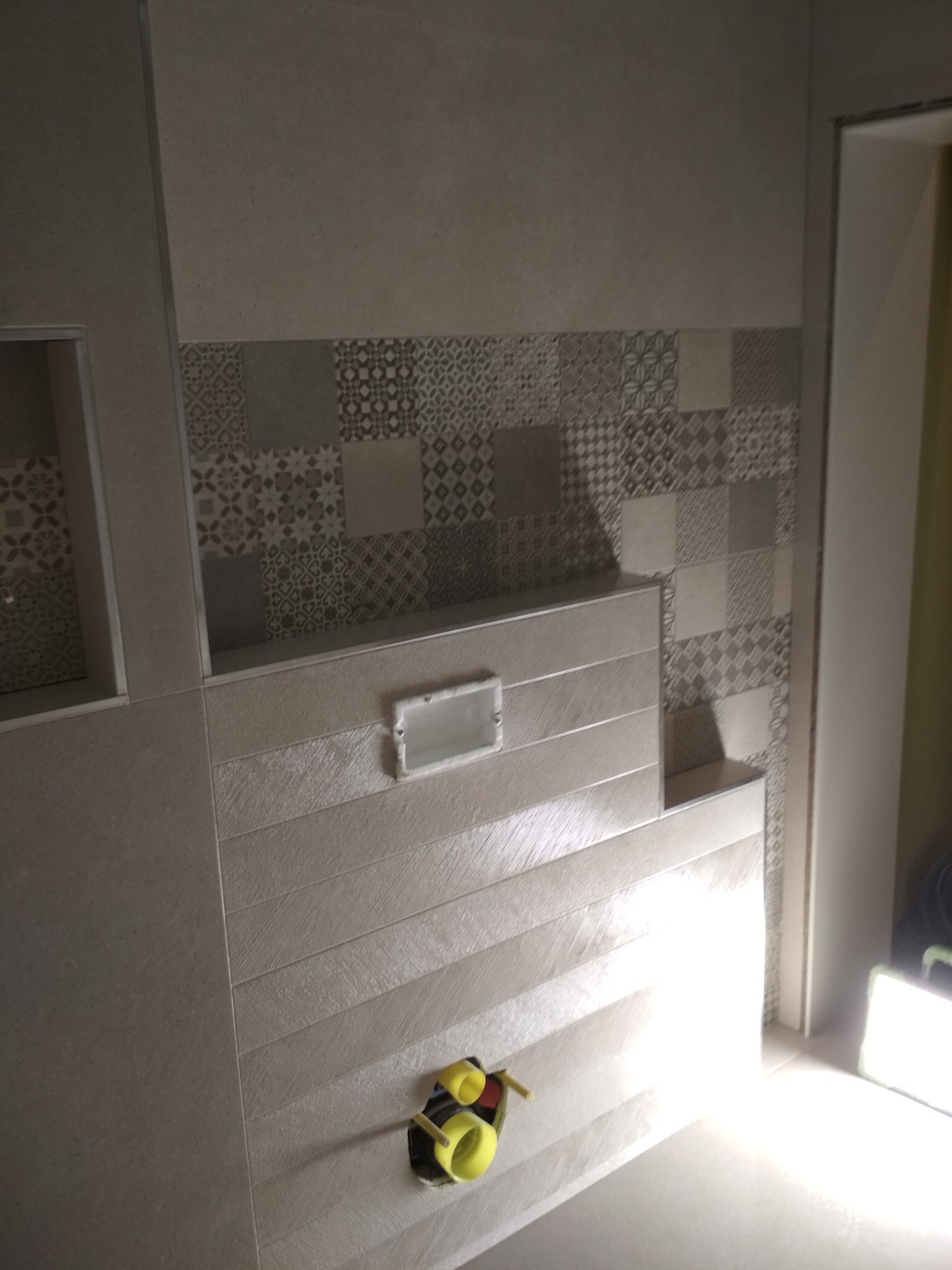 Fragment ściany łazienki z ułożonymi płytkami w dwóch stylach: gładkie duże płyty oraz dekoracyjne mozaiki u góry, widoczne rury instalacji wodnej.