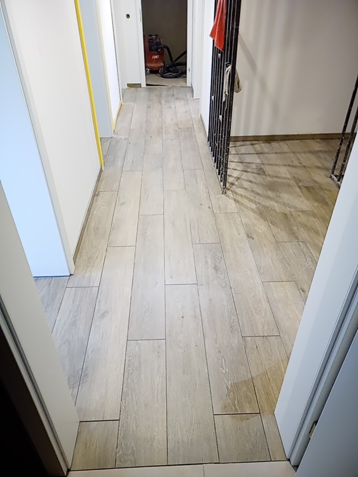 Korytarz z nową, jasną podłogą z płytek imitujących drewno, widoczna odkurzacz przemysłowy w tle i zabezpieczone żółtą taśmą malarską ściany.
