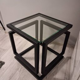 USŁUGI SPAWALNICZE I KONSERWACYJNE PATRYK WZGARDA - Nowoczesny stolik kawowy z czarną, metalową podstawą w kształcie sześcianu i szklanym blatem. Minimalistyczny design, widoczne łączenia spawane.