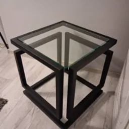 Nowoczesny stolik kawowy z czarną, metalową podstawą w kształcie sześcianu i szklanym blatem. Minimalistyczny design, widoczne łączenia spawane.