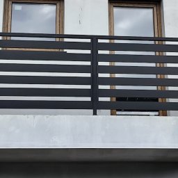 USŁUGI SPAWALNICZE I KONSERWACYJNE PATRYK WZGARDA - Czarna, pozioma, metalowa konstrukcja balkonu z widocznymi oknami w tle, prezentująca nowoczesny design i minimalistyczne wykończenie.