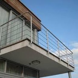 USŁUGI SPAWALNICZE I KONSERWACYJNE PATRYK WZGARDA - Nowoczesna balustrada ze stali nierdzewnej na balkonie budynku z drewnianą elewacją, widziana pod kątem, na tle błękitnego nieba z delikatnymi chmurami.