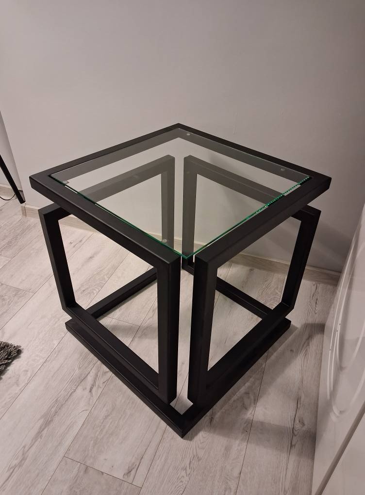 Nowoczesny stolik kawowy z czarną, metalową podstawą w kształcie sześcianu i szklanym blatem. Minimalistyczny design, widoczne łączenia spawane.