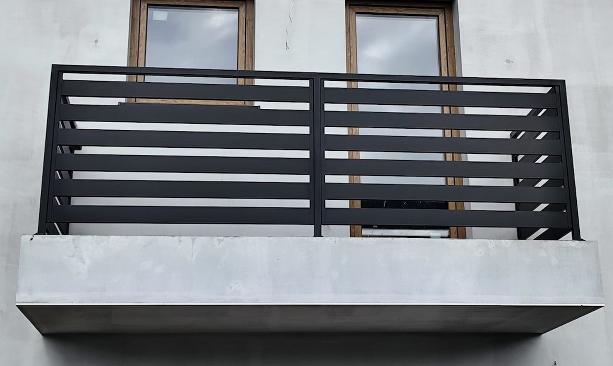 Czarna, pozioma, metalowa konstrukcja balkonu z widocznymi oknami w tle, prezentująca nowoczesny design i minimalistyczne wykończenie.