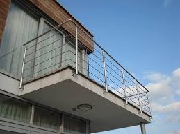 Nowoczesna balustrada ze stali nierdzewnej na balkonie budynku z drewnianą elewacją, widziana pod kątem, na tle błękitnego nieba z delikatnymi chmurami.