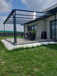 Nowoczesna pergola z czarną konstrukcją i przezroczystym zadaszeniem, przylegająca do jasnego budynku. Widoczny fragment ogrodu i meble ogrodowe.