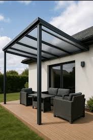 Nowoczesna pergola z grafitową konstrukcją i przezroczystym zadaszeniem, osłaniająca taras z meblami wypoczynkowymi przy białym budynku. Ujęcie w słoneczny dzień.