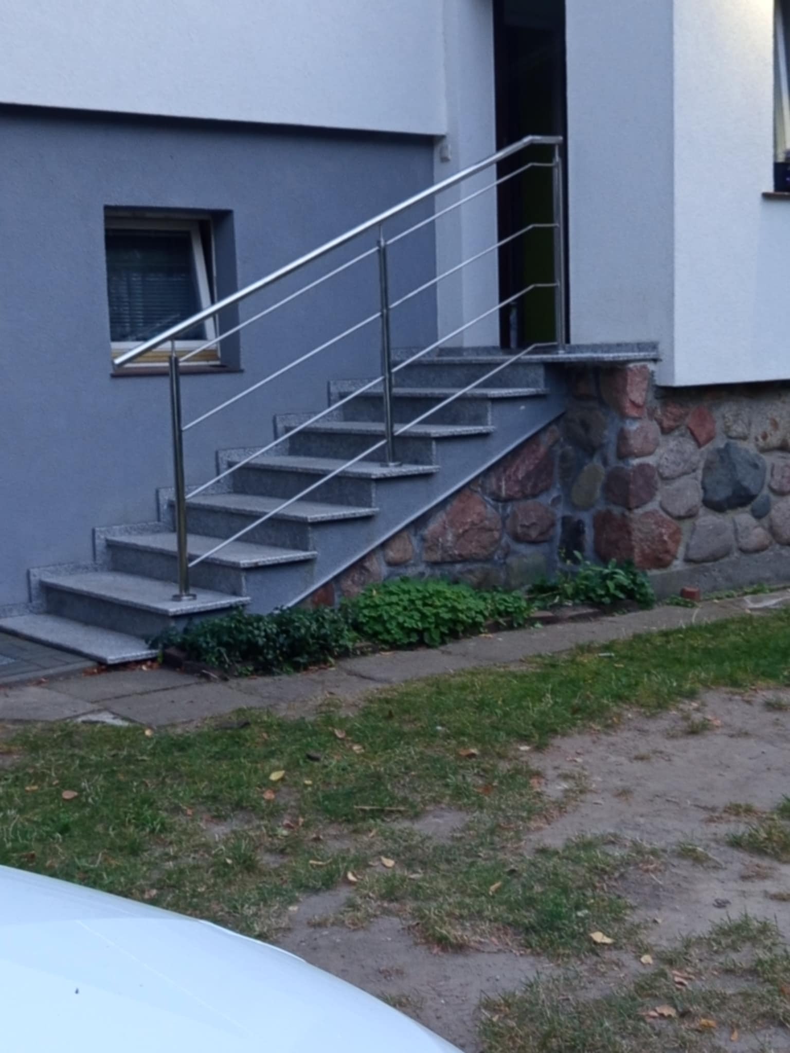 Zewnętrzne schody z szarego kamienia prowadzą do drzwi wejściowych, zabezpieczone balustradą ze stali nierdzewnej. Kamienna podmurówka budynku. Ujęcie z dołu.