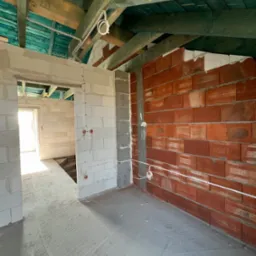 Wnętrze w stanie surowym: instalacja elektryczna natynkowa z puszkami na ścianach z cegły i betonu komórkowego, widoczne kable pod sufitem z drewnianą konstrukcją.