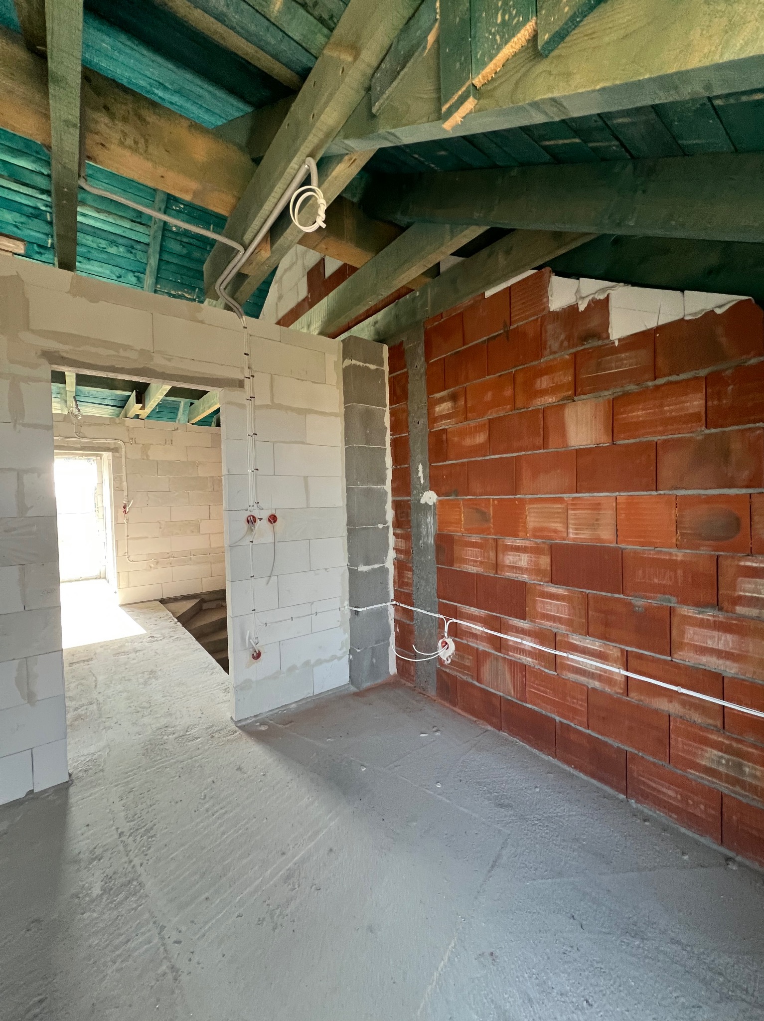 Wnętrze w stanie surowym: instalacja elektryczna natynkowa z puszkami na ścianach z cegły i betonu komórkowego, widoczne kable pod sufitem z drewnianą konstrukcją.