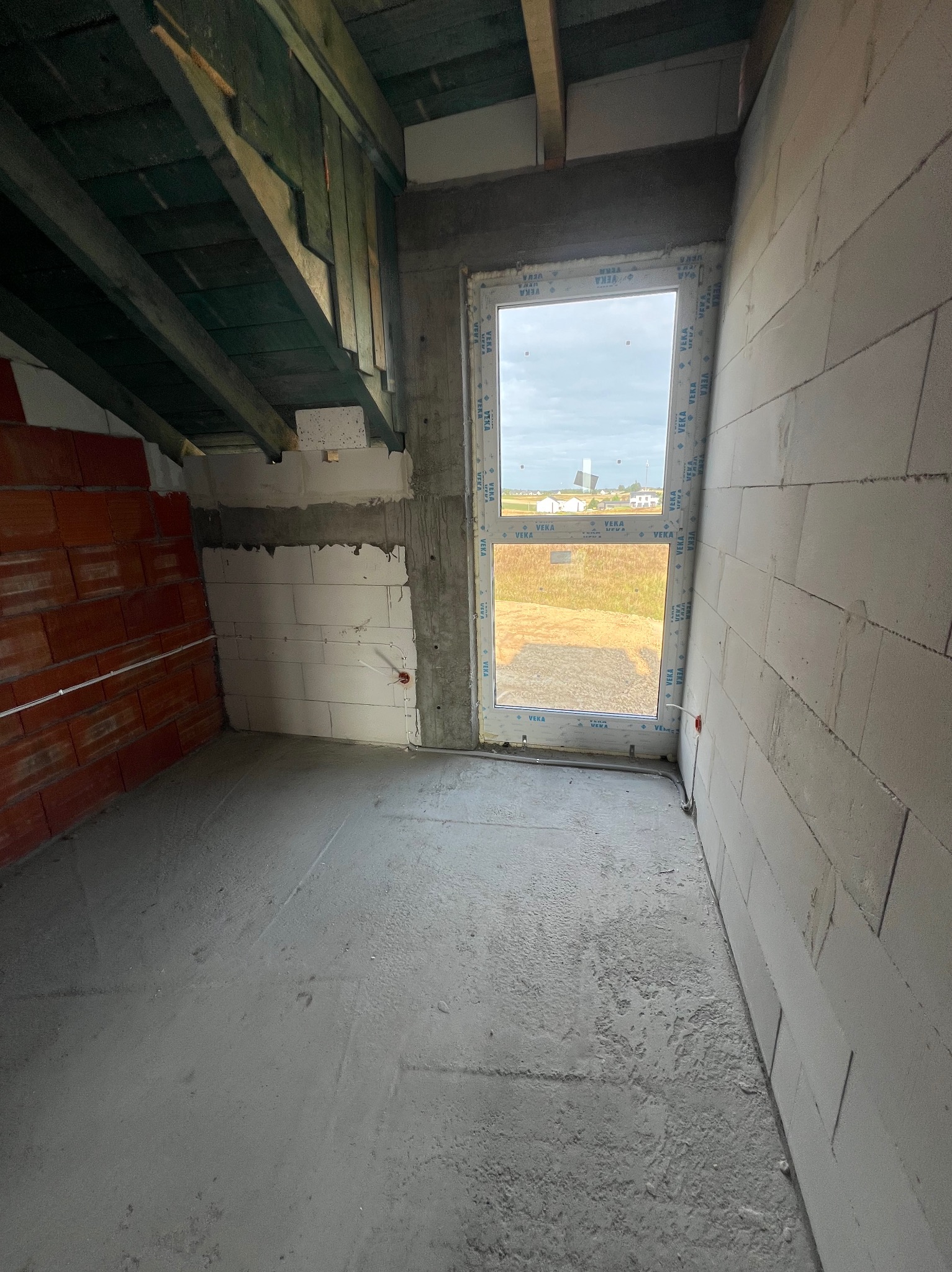 Surowy stan poddasza: ściany z bloczków i cegły, okno z widokiem na krajobraz, betonowa posadzka, widoczne przewody elektryczne. Elementy instalacji elektrycznej.