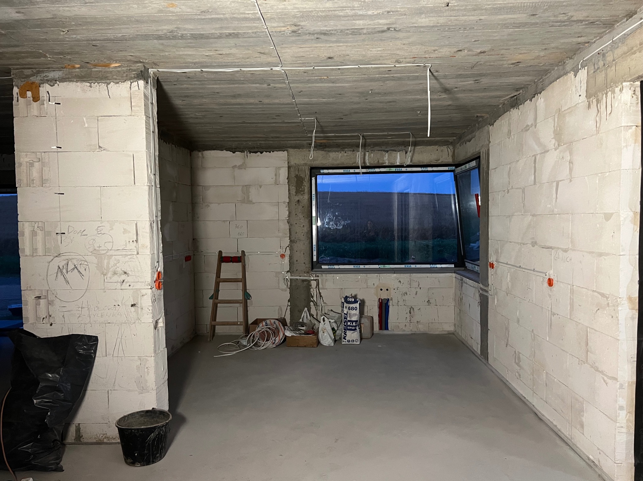 Surowy stan wnętrza z widocznymi kablami elektrycznymi pod sufitem, ściany z betonu komórkowego, okno z widokiem na zewnątrz i narzędzia na podłodze.