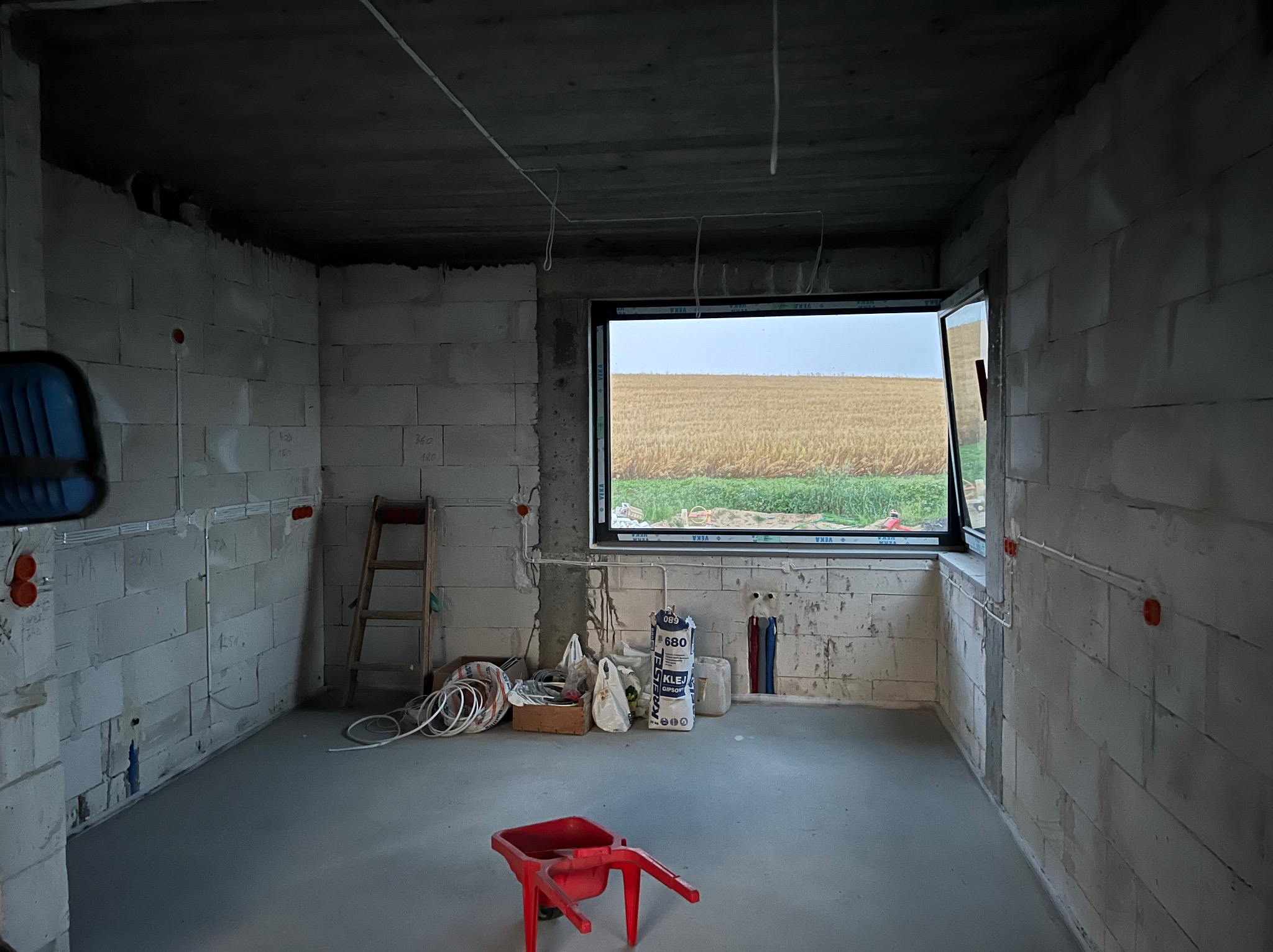 Surowy stan wnętrza z widoczną instalacją elektryczną (puszki, przewody) na ścianach z betonu komórkowego. W tle okno z widokiem na pole. Czerwony wózek na pierwszym planie.