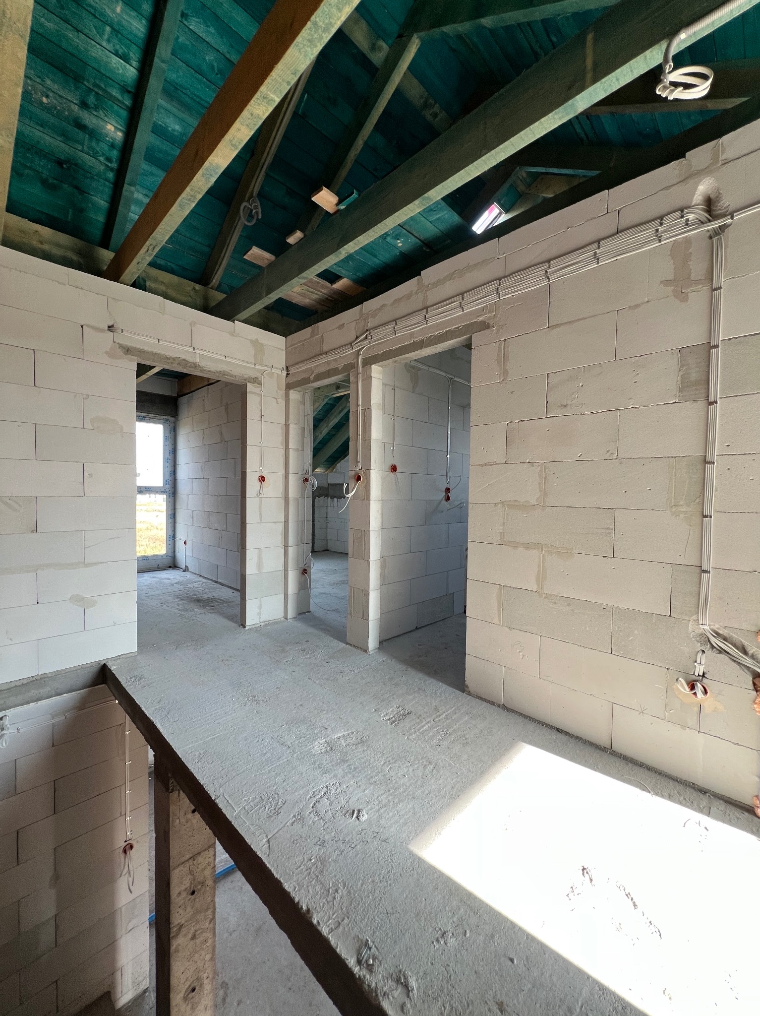 Wnętrze w stanie surowym z widoczną instalacją elektryczną: kable w peszlach przytwierdzone do ściany z betonu komórkowego, sufit z drewnianych belek pomalowanych na niebiesko.