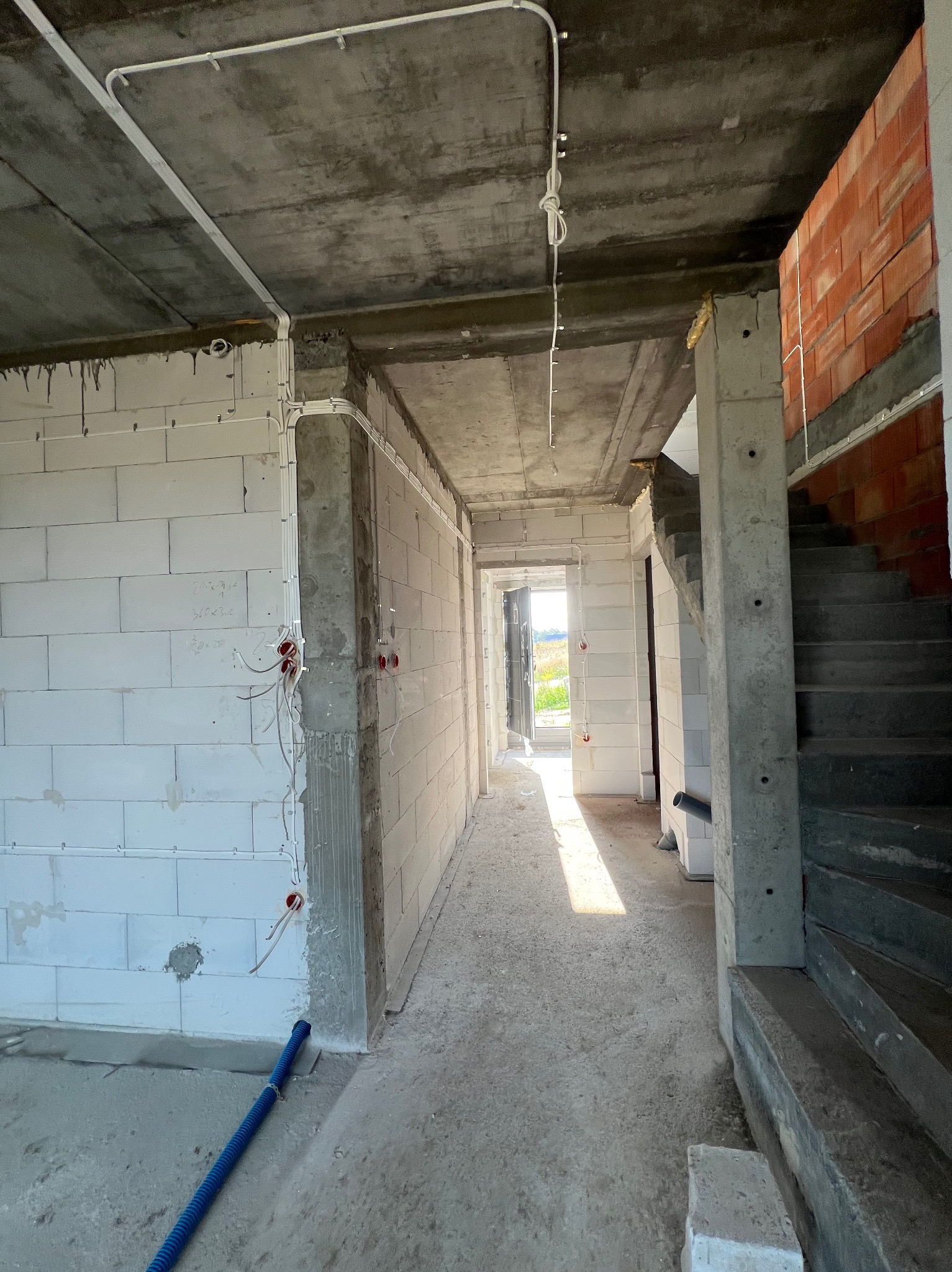 Wnętrze w stanie surowym z widoczną instalacją elektryczną: kable poprowadzone po ścianach i suficie, puszki elektryczne w ścianach. Widoczne schody i fragment muru z cegły.