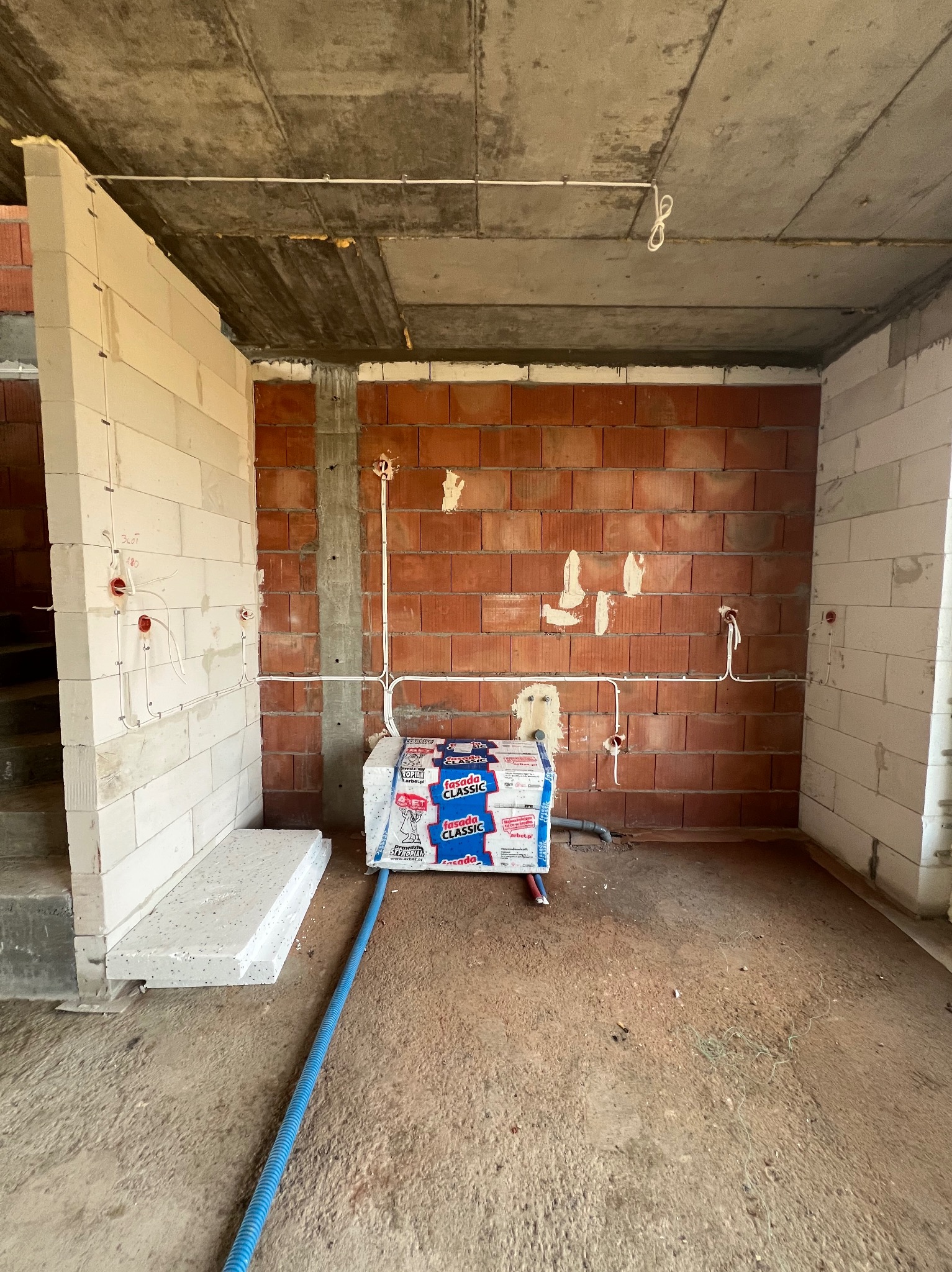 Surowy stan: białe i czerwone cegły, przewody elektryczne w ścianach. Instalacja w trakcie budowy, widoczne materiały budowlane.