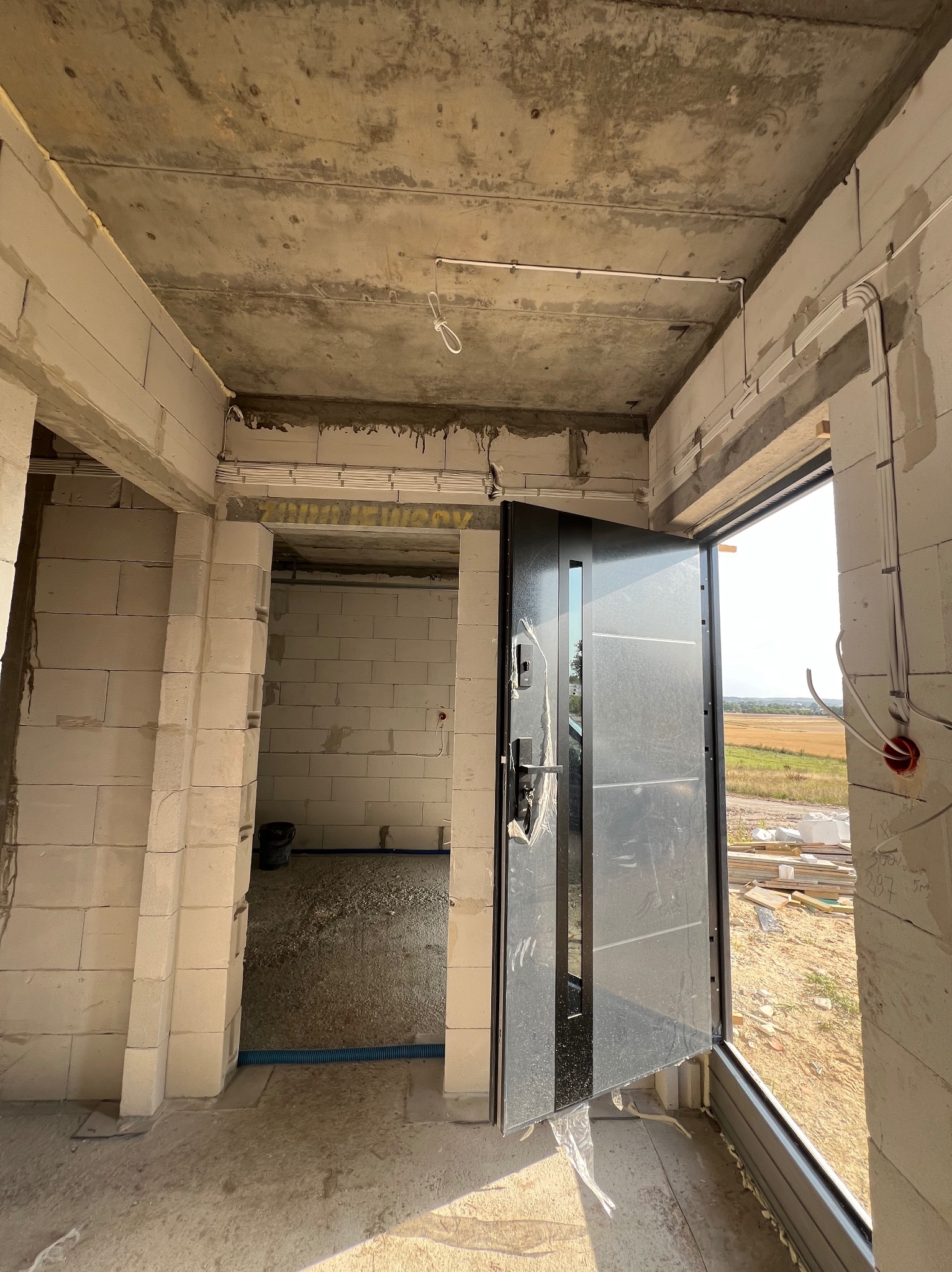 Surowy stan: wnętrze w trakcie budowy z widocznymi przewodami elektrycznymi na suficie, otwartymi drzwiami wejściowymi i pustakami.