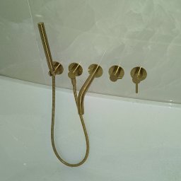 HMS instalacje hydrauliczne Marcin Szymański - Złota bateria wannowa ścienna z deszczownicą i czterema pokrętłami, zamontowana nad białą wanną z widoczną etykietą producenta Cersanit.