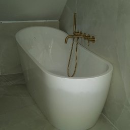 HMS instalacje hydrauliczne Marcin Szymański - Wolnostojąca wanna z baterią w kolorze złotym, zamontowana w łazience wykończonej płytkami imitującymi marmur, widok z boku i góry, oświetlenie górne.