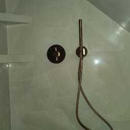 HMS instalacje hydrauliczne Marcin Szymański - Nowoczesna kabina prysznicowa z marmurowymi płytkami, wyposażona w złotą baterię termostatyczną i słuchawkę prysznicową na wężu, zamontowaną na ścianie.