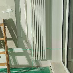 HMS instalacje hydrauliczne Marcin Szymański - Biały grzejnik panelowy zamontowany na ścianie, wyznaczony poziom lasera, drabina, narzędzia na podłodze zabezpieczonej folią malarską, widoczne elementy dekoracyjne ścian.