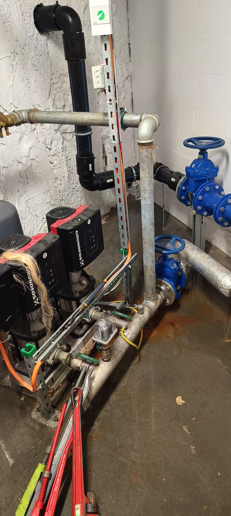 Piwnica z instalacją hydrauliczną: dwie pompy Grundfos, rury stalowe i plastikowe, zawory motylkowe w kolorze niebieskim, podłoga z zaciekami, klucze hydrauliczne na posadzce.