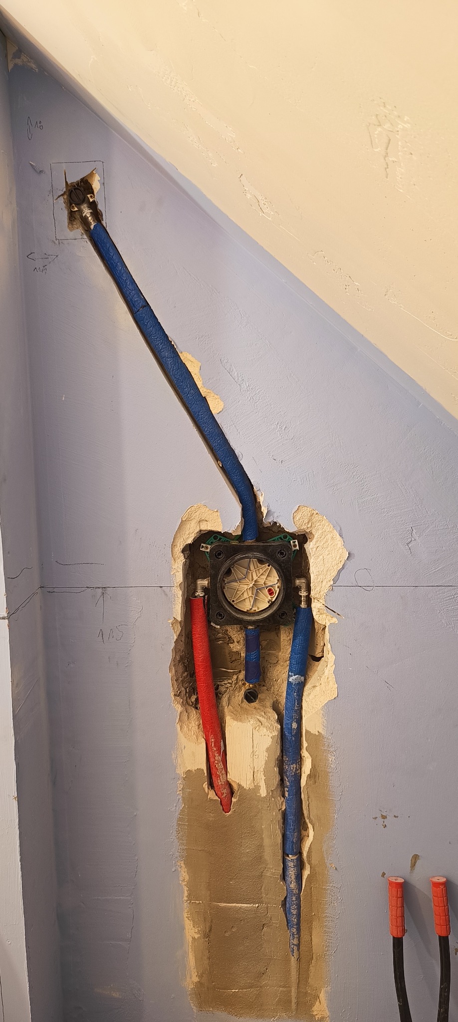 Wnęka w ścianie z widoczną instalacją hydrauliczną: rozdzielacz wody z podłączonymi rurami PEX w kolorach czerwonym i niebieskim, ściana w odcieniach fioletu i beżu z widocznymi śladami po pracach...