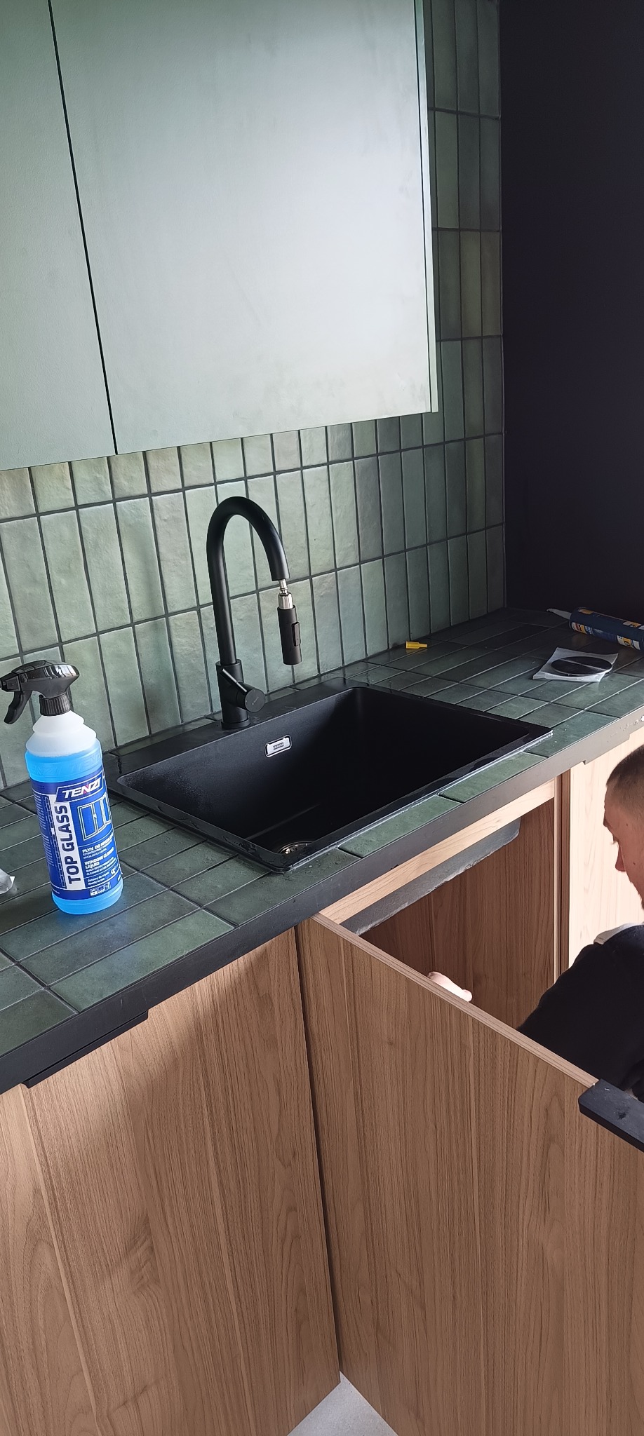Czarny zlew Franke z baterią z wyciąganą wylewką, butelka środka czyszczącego Tenzi Top Glass, zielone płytki na ścianie i blatach, montaż szafki zlewozmywakowej z drewnianymi frontami.