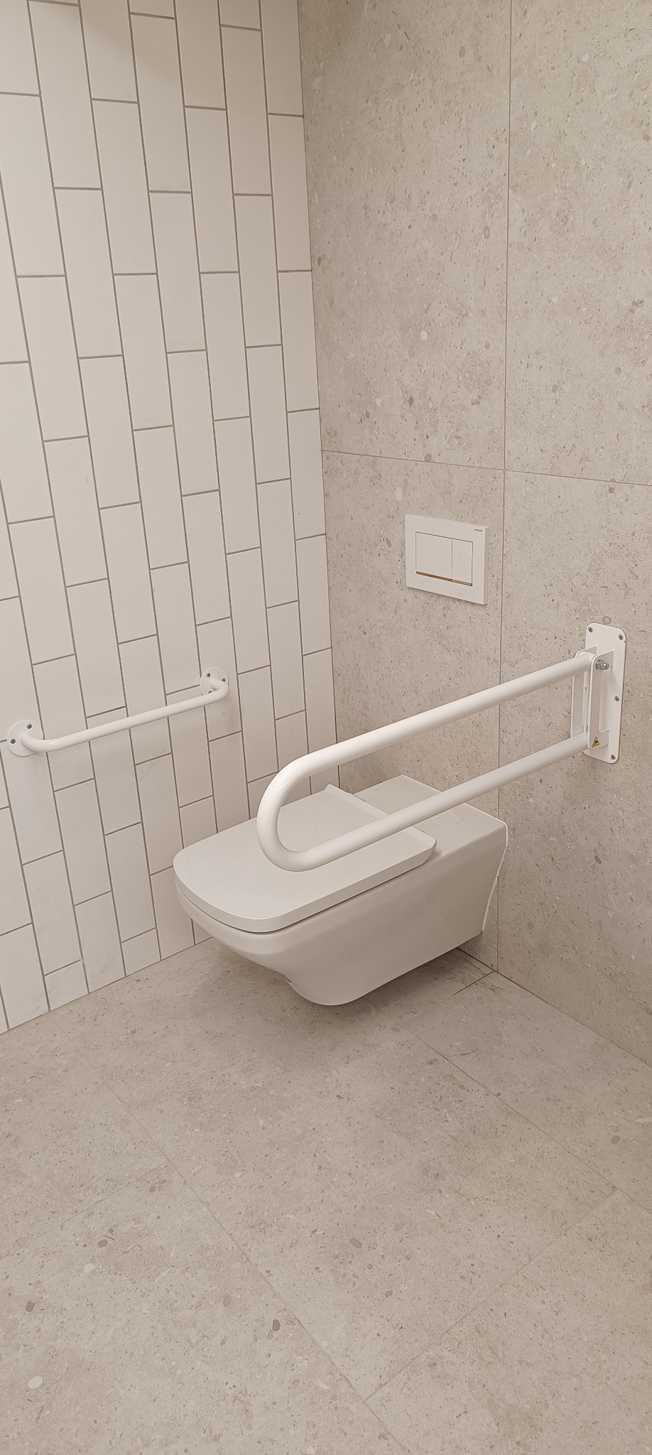 Podwieszana toaleta z uchwytami dla osób niepełnosprawnych zamontowana na ścianie wyłożonej płytkami imitującymi kamień, obok ściana wyłożona białymi, pionowymi płytkami.