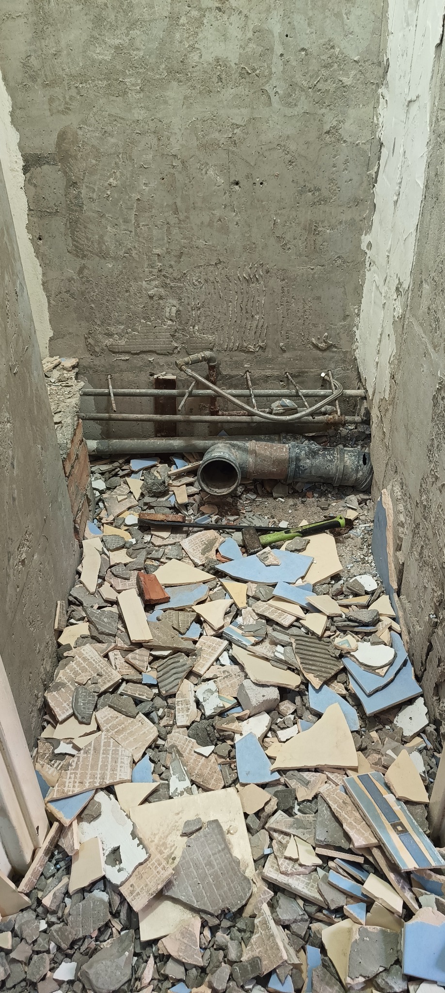 Widok z góry na fragment instalacji hydraulicznej w trakcie demontażu, z widocznymi rurami, gruzem i fragmentami potłuczonych płytek w pomieszczeniu o surowych, betonowych ścianach.