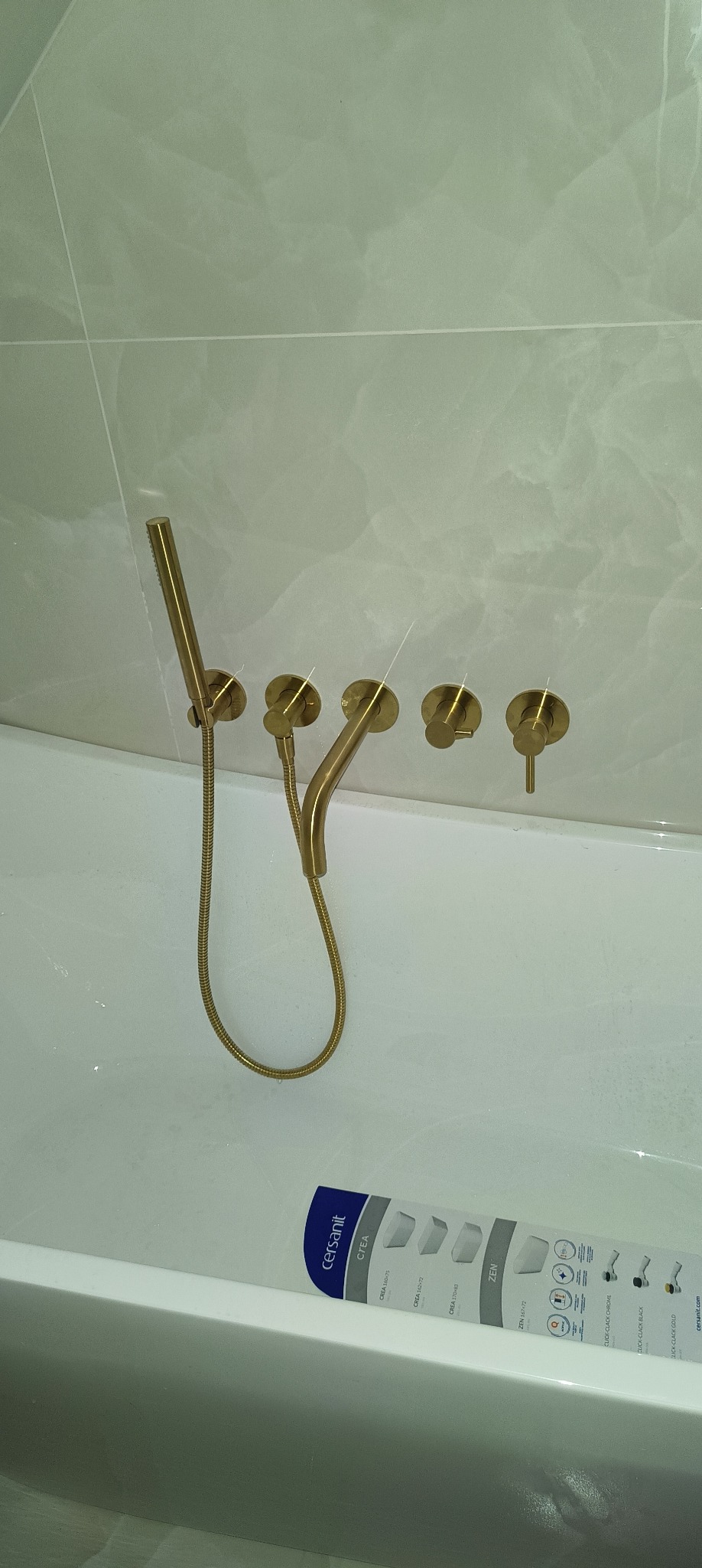 Złota bateria wannowa ścienna z deszczownicą i czterema pokrętłami, zamontowana nad białą wanną z widoczną etykietą producenta Cersanit.