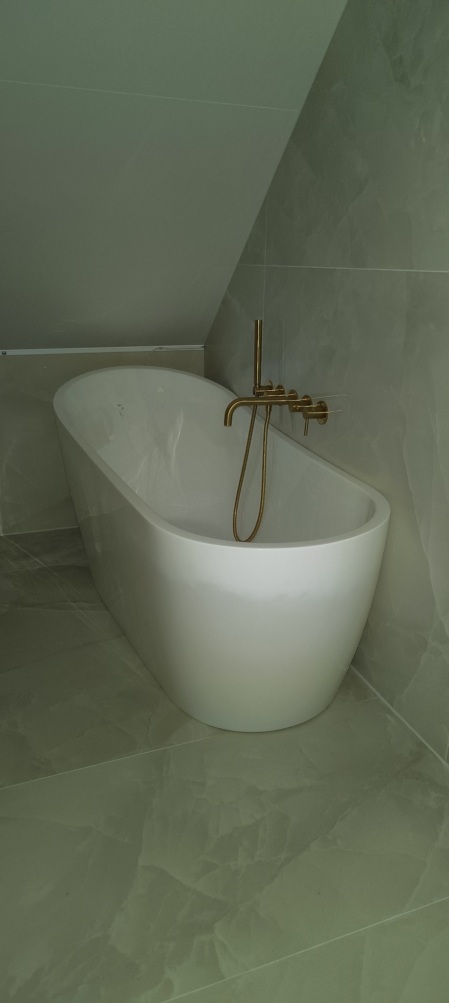 Wolnostojąca wanna z baterią w kolorze złotym, zamontowana w łazience wykończonej płytkami imitującymi marmur, widok z boku i góry, oświetlenie górne.