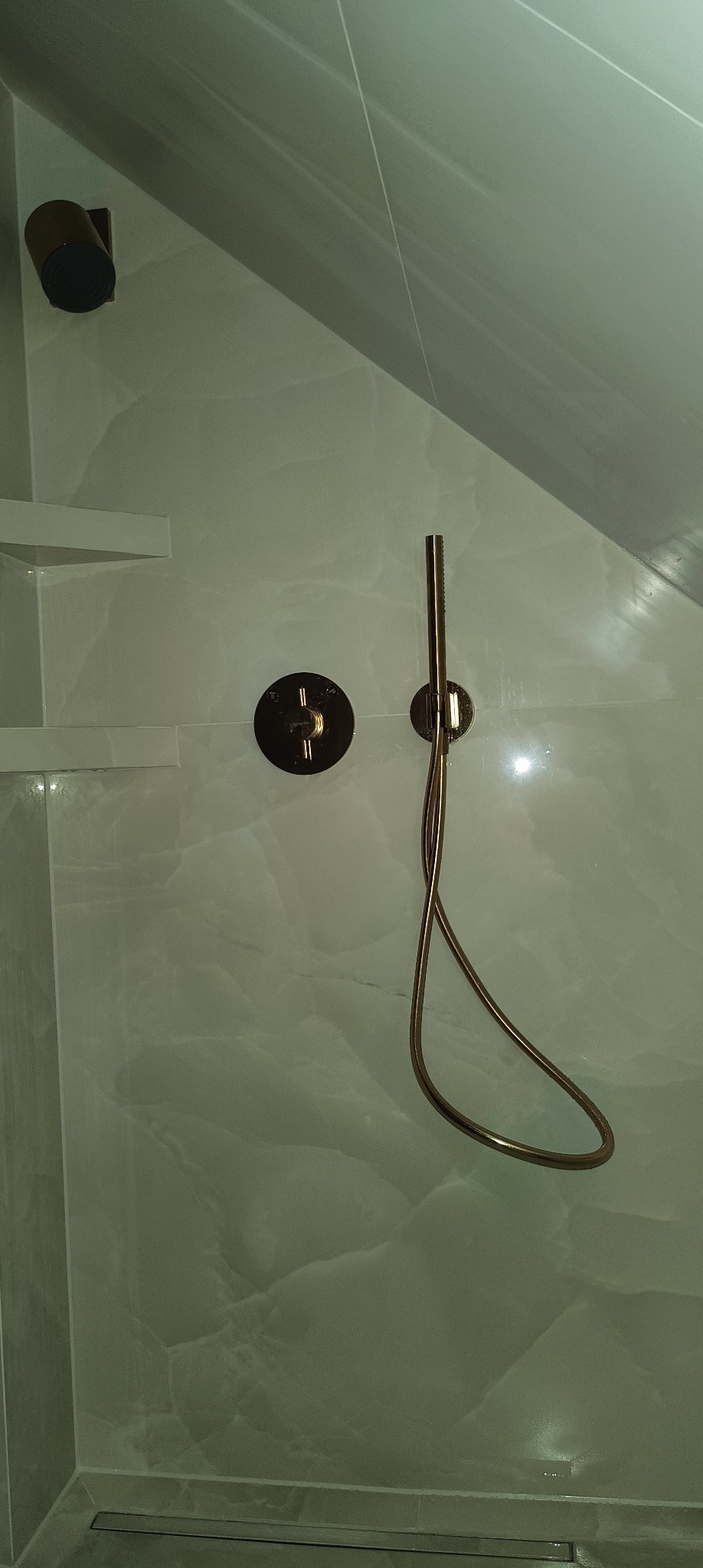 Nowoczesna kabina prysznicowa z marmurowymi płytkami, wyposażona w złotą baterię termostatyczną i słuchawkę prysznicową na wężu, zamontowaną na ścianie.