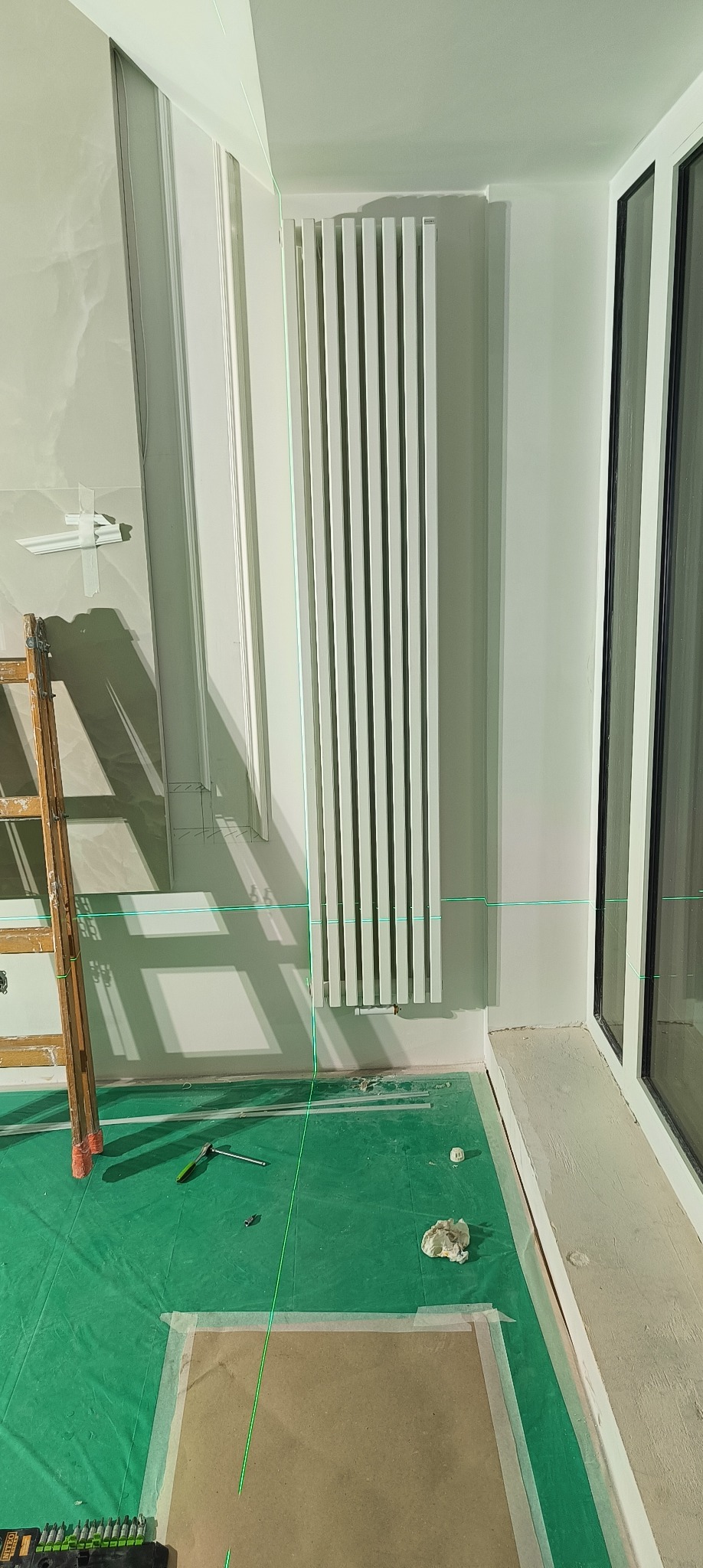 Biały grzejnik panelowy zamontowany na ścianie, wyznaczony poziom lasera, drabina, narzędzia na podłodze zabezpieczonej folią malarską, widoczne elementy dekoracyjne ścian.