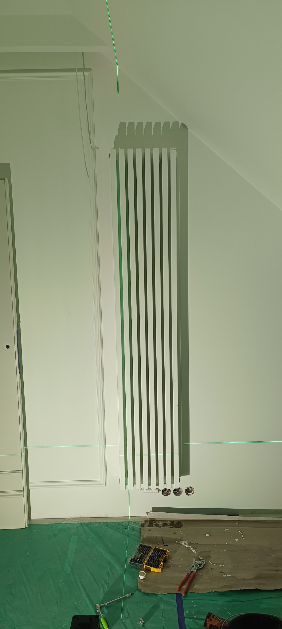 Biały grzejnik panelowy zawieszony na ścianie, z widocznymi otworami przyłączeniowymi, poziomica laserowa wyznacza linię, narzędzia i materiały robocze na podłodze zabezpieczonej folią.