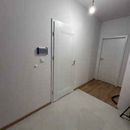 ADEOBUD - Białe drzwi wewnętrzne z złotą klamką, częściowo otwarte, na tle białych ścian i jasnej podłogi. Widoczny fragment drewnianej podłogi w sąsiednim pomieszczeniu.