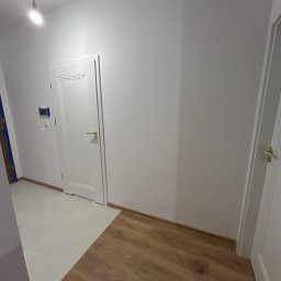 ADEOBUD - Białe drzwi z złotą klamką w nowym wnętrzu. Podłoga łączy panele w odcieniach brązu z jasnymi płytkami. Ściany pomalowane na biało. Widoczny fragment odkurzacza.