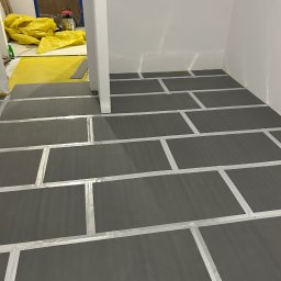 ADEOBUD - Przygotowanie podłoża: ciemne płyty podkładowe ułożone na podłodze, zabezpieczone taśmą, gotowe do dalszych prac wykończeniowych. W tle folia ochronna i sprzęt.