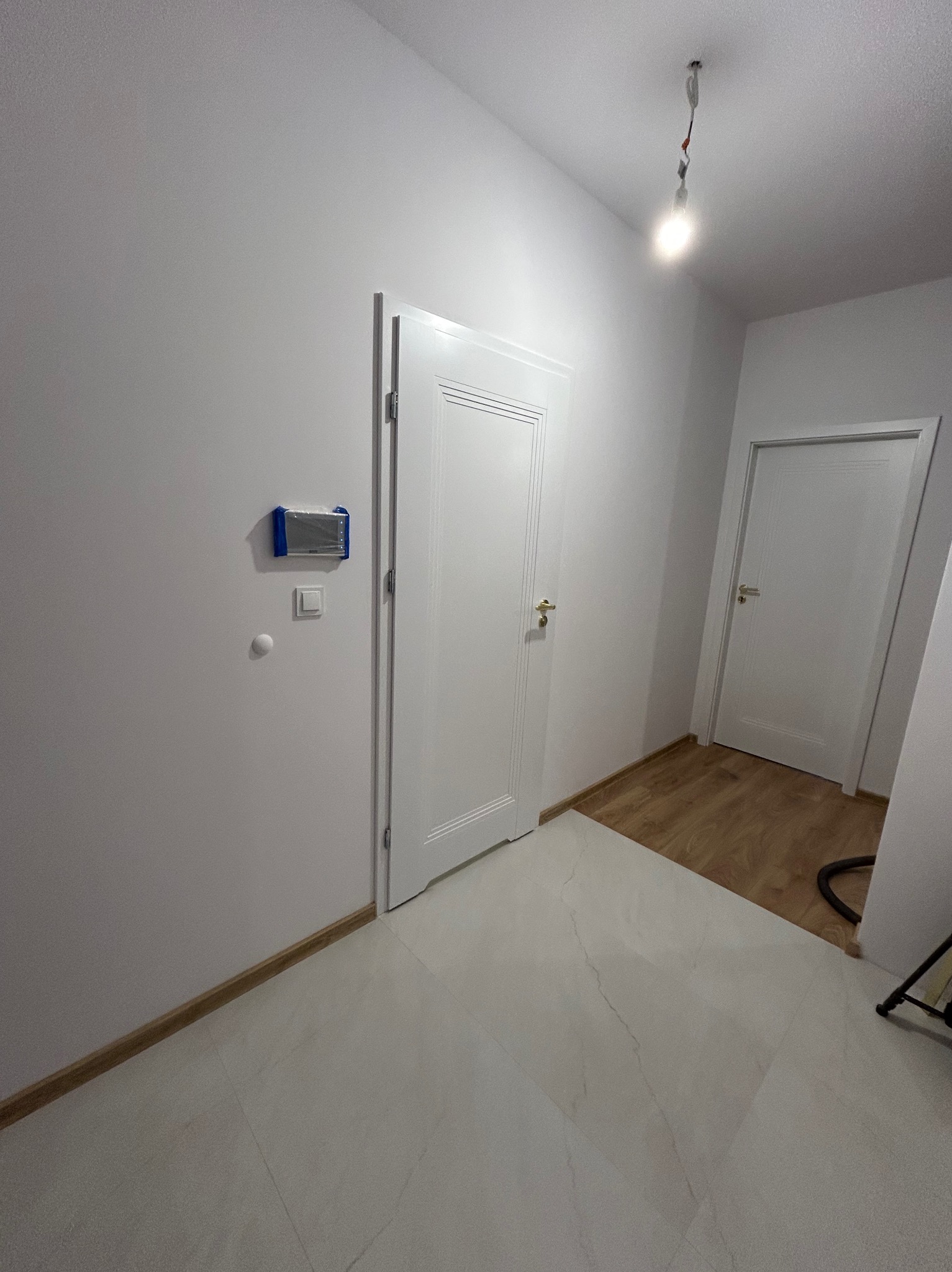 Białe drzwi wewnętrzne z złotą klamką, częściowo otwarte, na tle białych ścian i jasnej podłogi. Widoczny fragment drewnianej podłogi w sąsiednim pomieszczeniu.