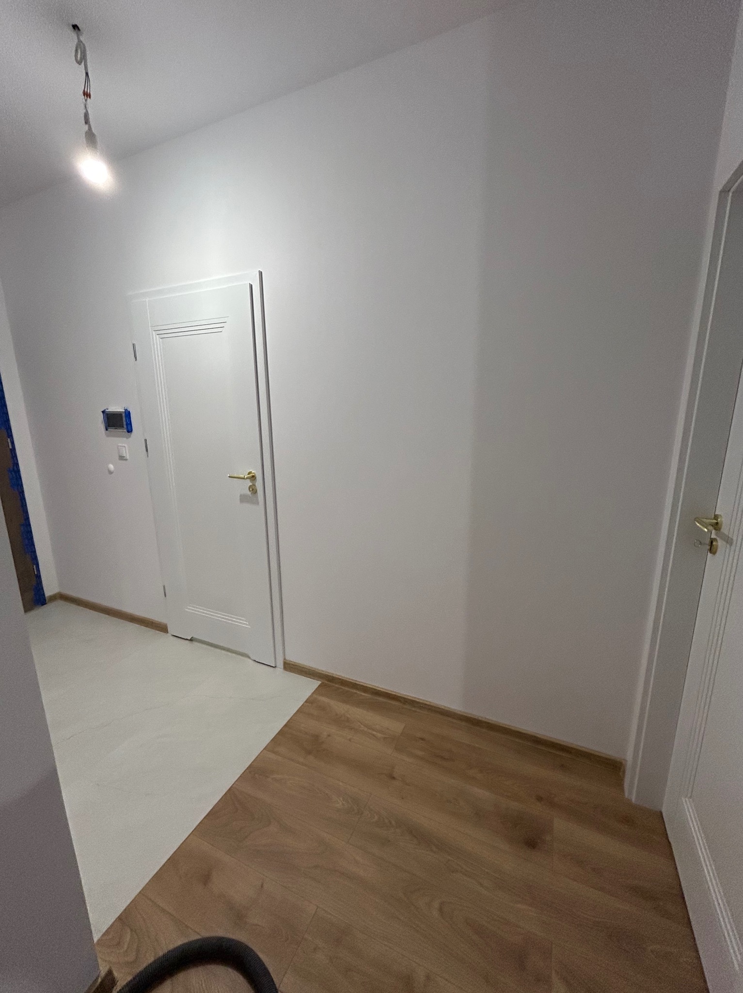 Białe drzwi z złotą klamką w nowym wnętrzu. Podłoga łączy panele w odcieniach brązu z jasnymi płytkami. Ściany pomalowane na biało. Widoczny fragment odkurzacza.