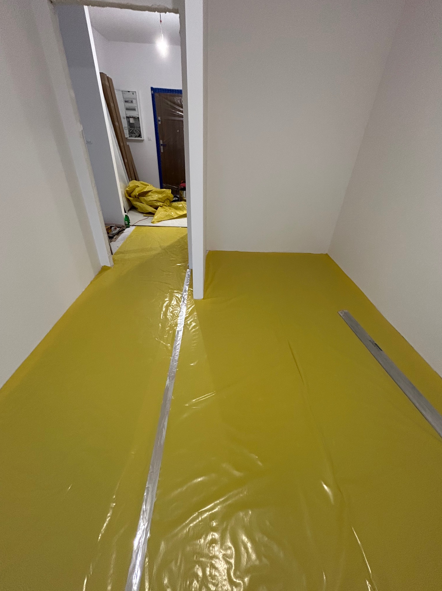 Przygotowanie podłoża: żółta folia ochronna rozłożona na podłodze w pustym pomieszczeniu, listwa przypodłogowa w rogu. Widok na drzwi wejściowe.
