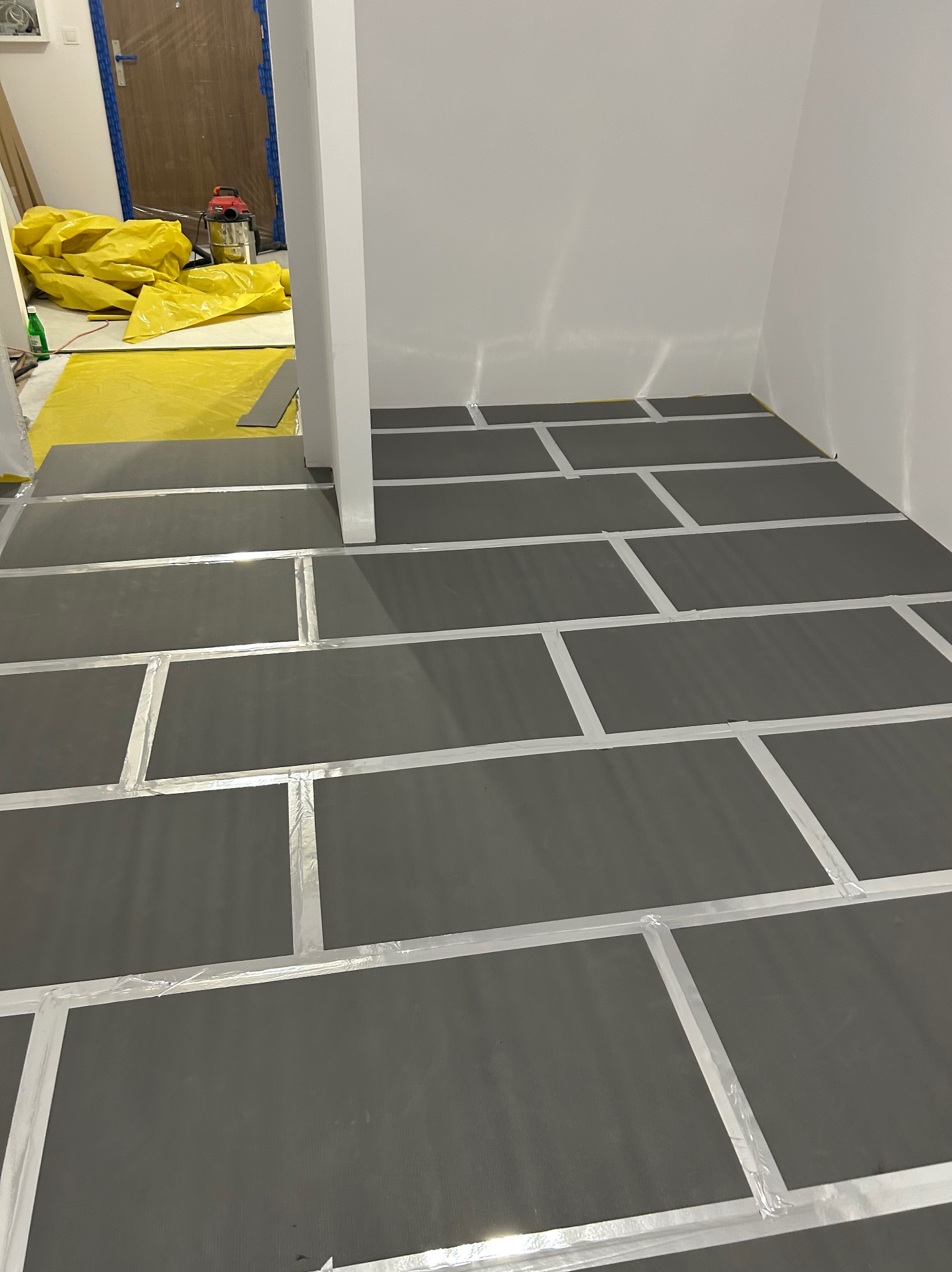 Przygotowanie podłoża: ciemne płyty podkładowe ułożone na podłodze, zabezpieczone taśmą, gotowe do dalszych prac wykończeniowych. W tle folia ochronna i sprzęt.