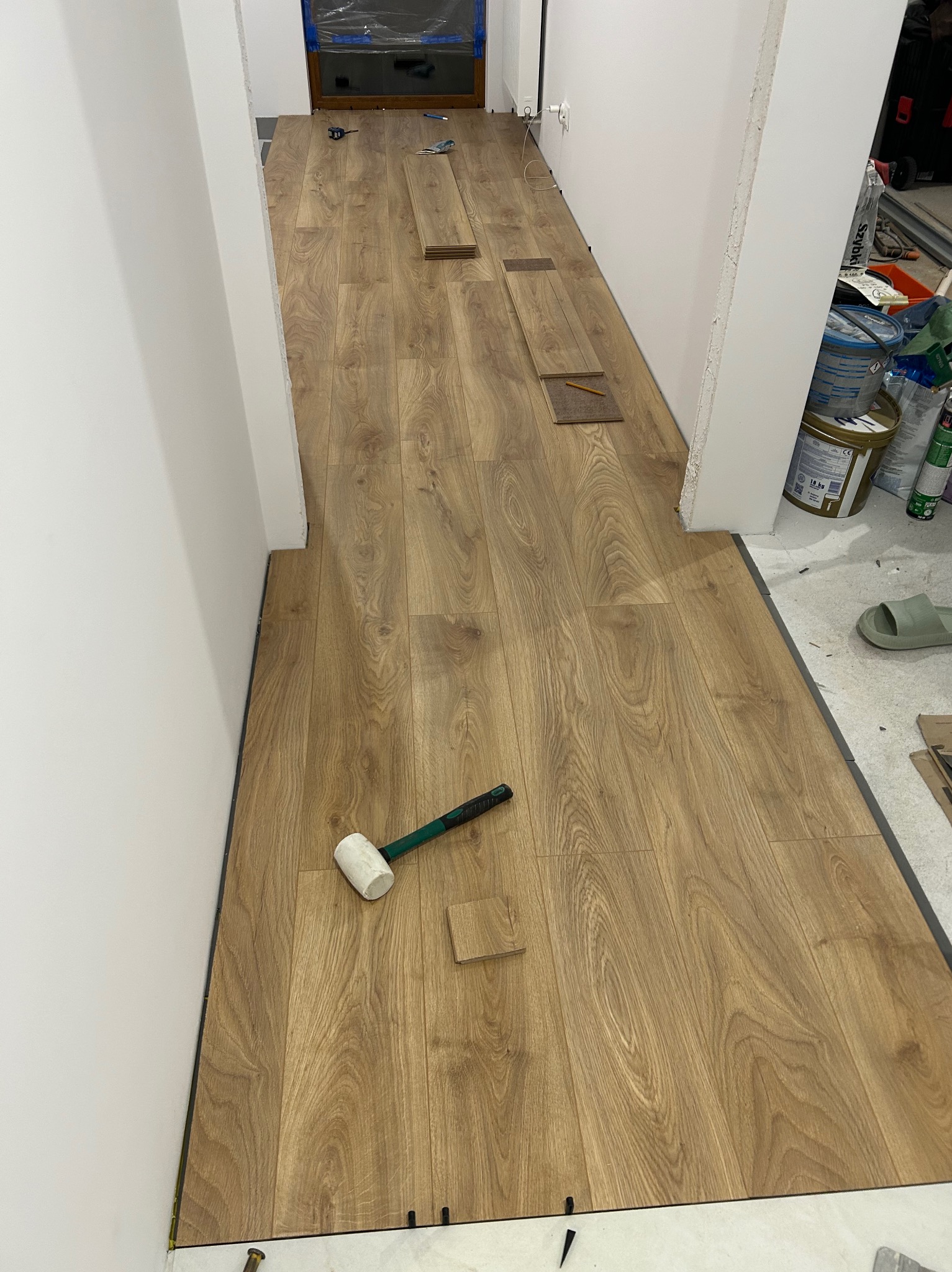 Układanie paneli podłogowych w korytarzu, widoczne narzędzia i fragmenty nieułożonych paneli. Jasne ściany, ciepłe odcienie drewna, etap prac w trakcie realizacji.