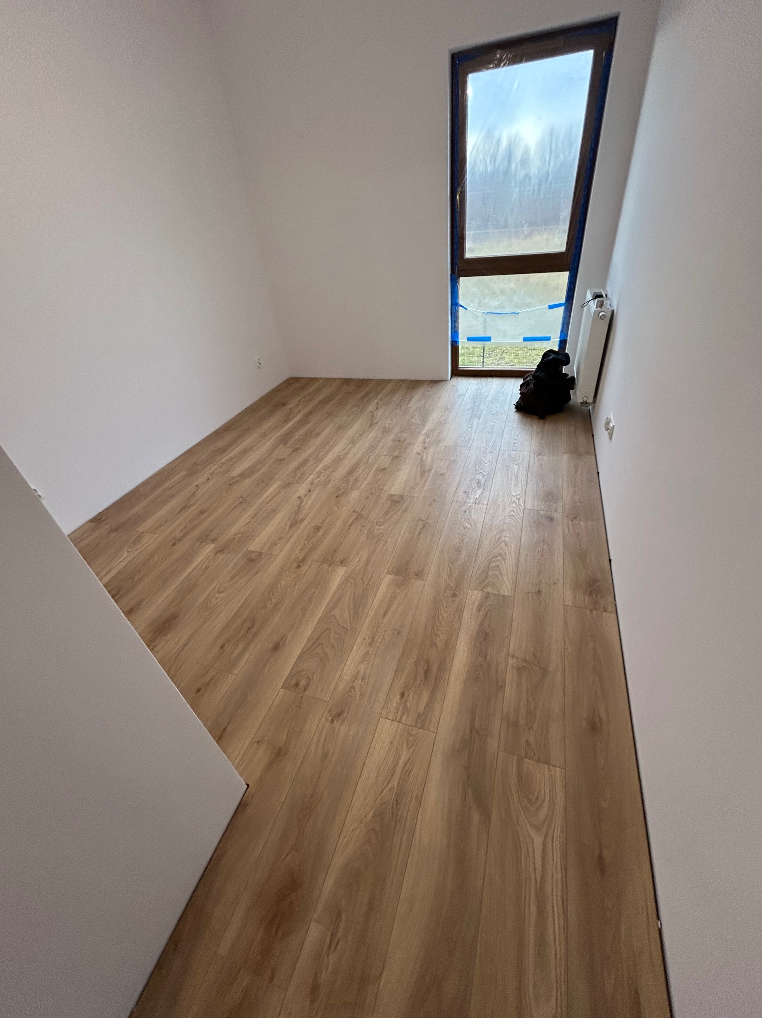 Jasne panele podłogowe ułożone w pokoju z oknem zabezpieczonym folią malarską. Ściany pomalowane na biało. Widoczny fragment ściany z płyty karton gips.