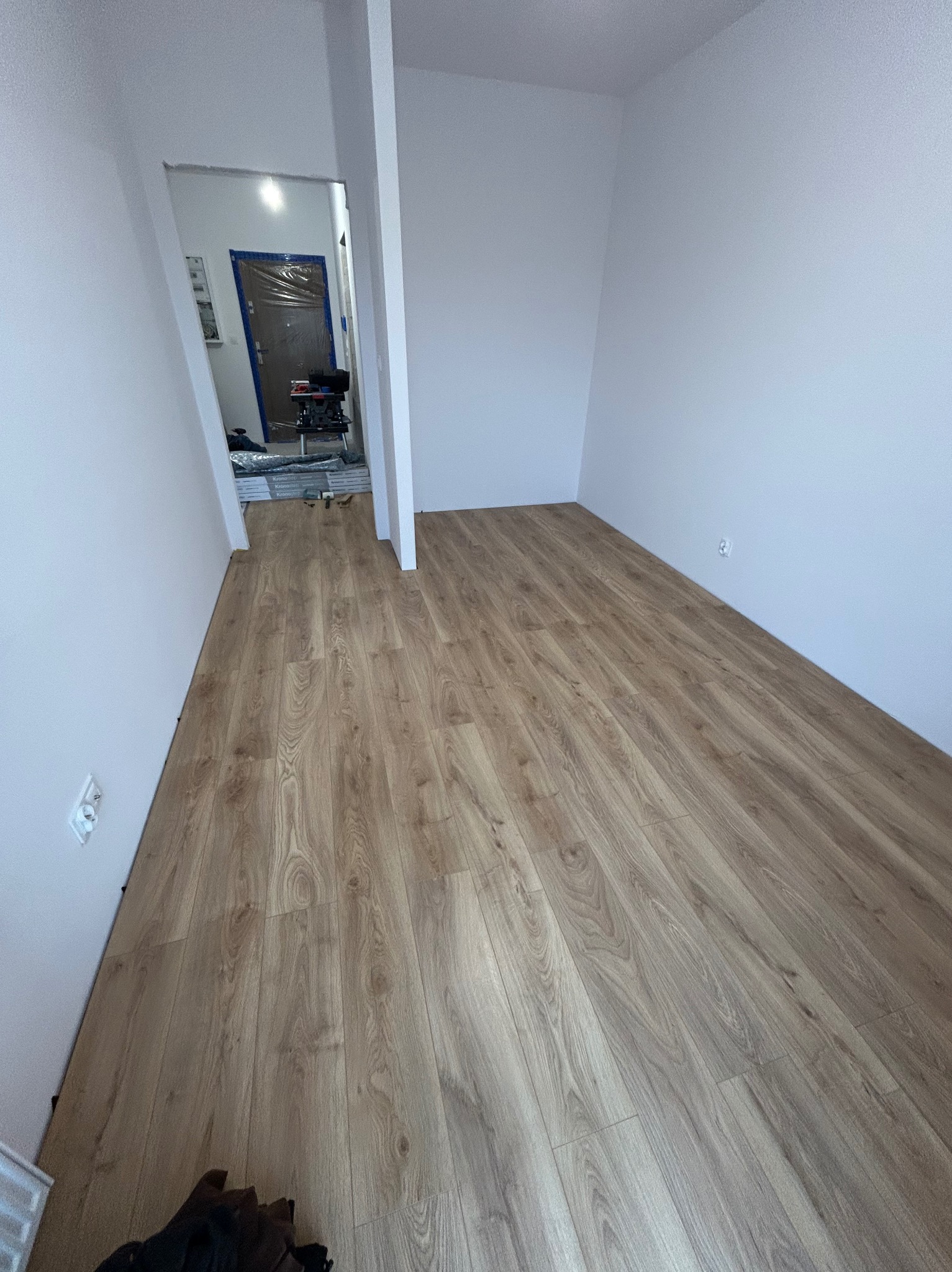 Jasne panele podłogowe ułożone w pokoju z białymi ścianami. W tle widoczne drzwi zabezpieczone folią malarską i narzędzia.