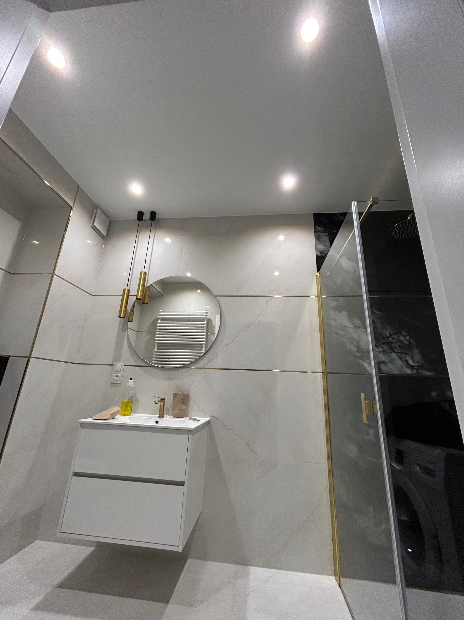 Nowoczesna łazienka z białymi płytkami, złotymi akcentami i okrągłym lustrem. Kabina prysznicowa z czarnym szkłem i złotymi okuciami. Minimalistyczna umywalka.