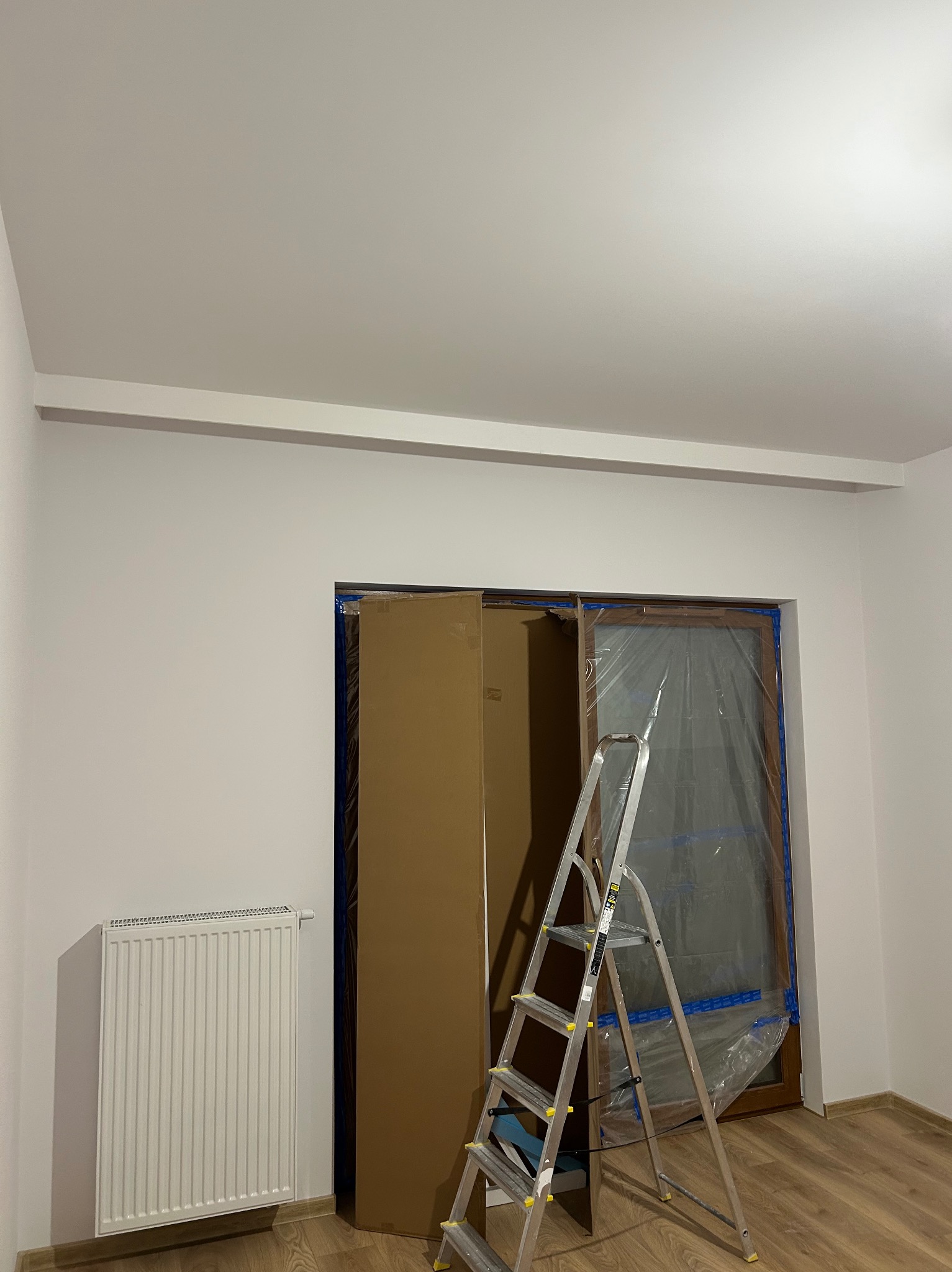Pokój w trakcie przygotowań do prac. Okno zabezpieczone folią, drabina, grzejnik i kartonowe zabezpieczenie drzwi. Ściany pomalowane na biało, podłoga drewniana.