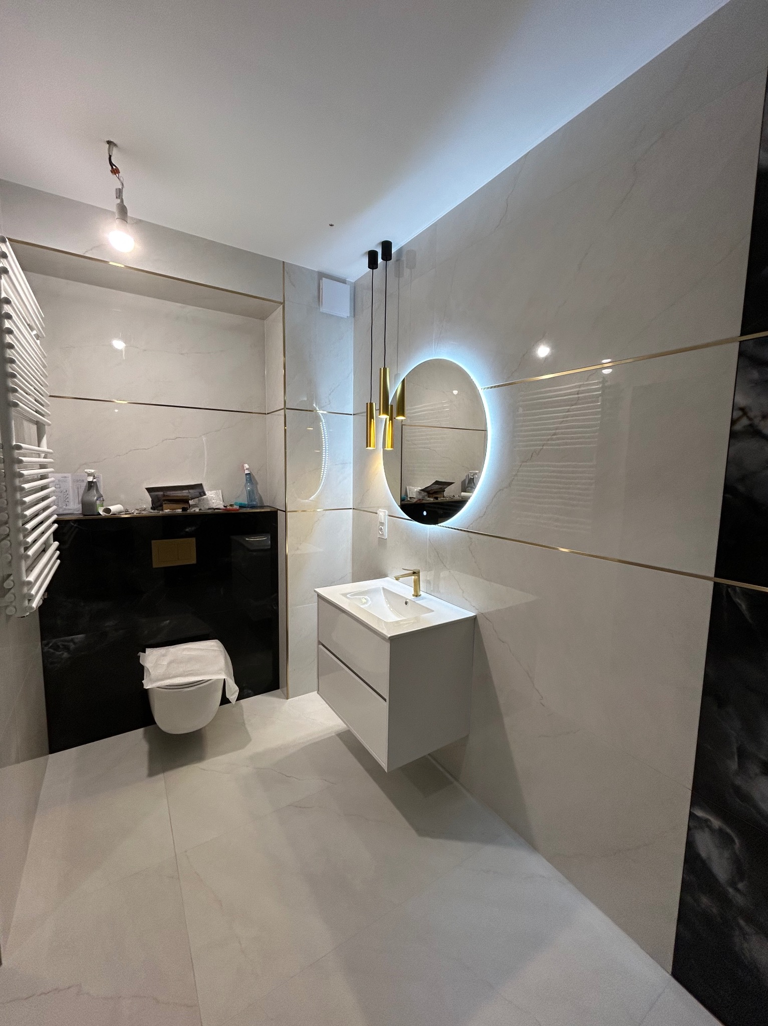 Nowoczesna łazienka z białymi płytkami imitującymi marmur, podświetlanym lustrem i złotymi akcentami. Uwagę zwraca wisząca toaleta i minimalistyczna umywalka.