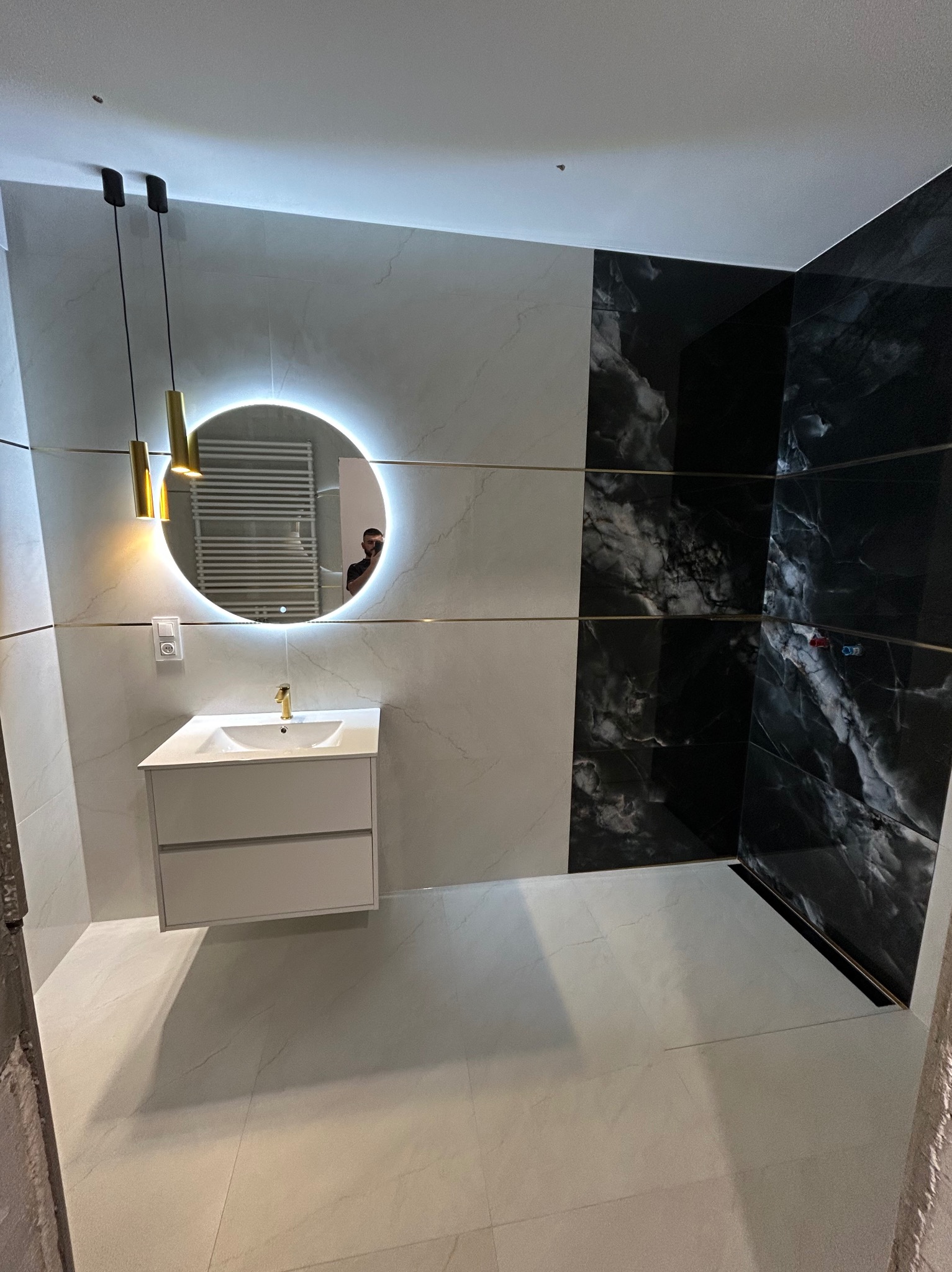 Wykończona łazienka z białymi i czarnymi płytkami imitującymi marmur, okrągłym lustrem z podświetleniem LED i wiszącą szafką z umywalką. Nowoczesny design.